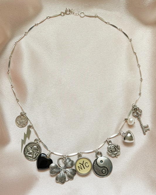 A silver charm necklace laid flat on a light beige satin fabric background. The necklace features multiple charms including a black heart, a silver clover, a round charm with 'NYC' in gold, a yin-yang symbol, a key, a pearl, and other small metallic icons.